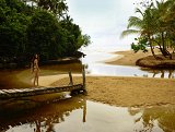 The image depicts a serene beach scene featuring a young woman standing on a wooden bridge that leads over a shallow stream. The setting includes lush greenery on either side, with tropical trees and plants. In the background, the sandy beach and ocean are visible under a cloudy sky. The overall mood is tranquil and picturesque, capturing the beauty of nature.