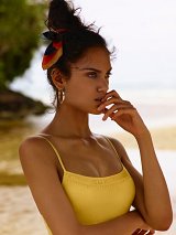 The image features a young woman standing on a beach. She has long, dark hair styled in a messy bun, adorned with a colorful scarf. She is wearing a yellow tank top that complements her skin tone. The background depicts a serene beach scene with soft waves and greenery, creating a tranquil atmosphere. The woman has a contemplative expression, with her hand gently touching her mouth, suggesting a moment of reflection.