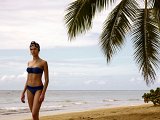 L'image montre une femme sur une plage, vêtue d'un maillot de bain bleu. Elle se tient près de l'eau, avec des palmiers en arrière-plan et un ciel légèrement nuageux. La scène évoque une atmosphère estivale et paisible, avec des couleurs naturelles et une ambiance relaxante.