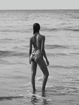 The image features a person standing in shallow water, facing away from the viewer. The scene is in black and white, emphasizing the serene atmosphere. The subject is wearing a bikini and has long hair that appears wet. The ocean waves gently lap at their legs, creating a calm and tranquil setting. The horizon in the background suggests a vast expanse of water and sky, contributing to the overall sense of peace.