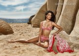 L'image présente une femme assise sur une plage de sable, vêtue d'un bikini rouge à motifs. Elle est élégante, avec les cheveux longs et sombres, regardant vers l'objectif avec une expression calme. À côté d'elle, un paréo assorti est drapé sur le sable. En arrière-plan, on peut voir l'océan scintillant sous le soleil et des rochers qui entourent la scène, créant une ambiance estivale et tranquille.