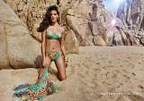The image features a woman posing on a sandy beach surrounded by large rock formations. She is wearing a colorful bikini with a vibrant, floral pattern. Her long, wavy hair flows elegantly, and she appears confident and poised. In one hand, she holds a flowing piece of fabric that adds a dynamic element to the scene. The backdrop showcases a sunny environment, with the sunlight creating a warm and inviting atmosphere. The bottom right corner displays the text "MARYAN MEHLHORN," suggesting a branding or designer label.