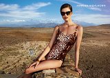 The image showcases a model posing in a stylish swimsuit with a leopard print design. She is wearing sunglasses and has a sophisticated demeanor. The background features a vast, arid landscape with mountains in the distance, creating a striking contrast with the model's casual yet elegant look. The lighting highlights the natural beauty of the setting and the design of the swimsuit. The brand name "MARYAN MEHLHORN" is displayed prominently in the corner.