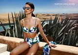 The image features a model posing in an outdoor setting. She is wearing a stylish bikini with a floral pattern, consisting of a top and high-waisted bottoms. The model has long, dark hair and is wearing sunglasses. In the background, there are desert-like sand dunes and lush green plants, which creates a striking contrast. The scene reflects a sunlit, warm atmosphere, suggesting a luxurious vacation vibe. The name "MARYAN MEHLHORN" is displayed in the corner, indicating a fashion brand.