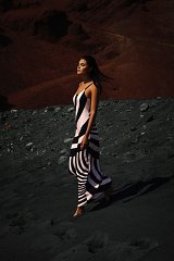 The image features a woman walking on a dark, textured surface, possibly sand or volcanic ash. She is wearing a long, form-fitting dress that has a bold, striped pattern with alternating colors, creating a striking visual contrast. The background consists of reddish-brown rocky formations, enhancing the dramatic atmosphere of the scene. The woman appears to be in a serene, contemplative pose, with her hair flowing gently in the breeze.